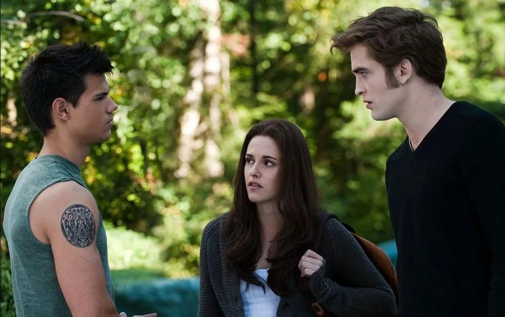 Da esquerda para a direita, Jacob, Bella e Edward aparecem em pé. Jacob e Edward se encaram com olhares sérios.