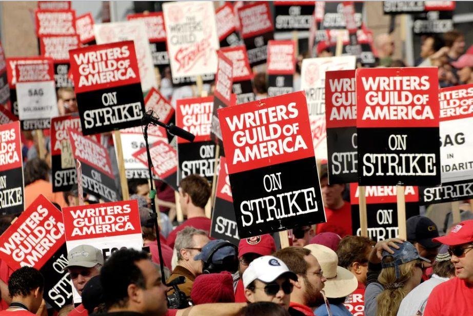 Diversos grevistas carregam cartazes preto e vermelho com os dizeres "Writers Guild of America on strike".