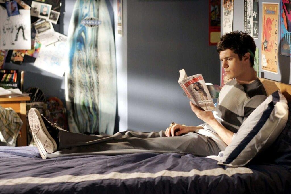 Seth Cohen (Adam Brody) aparece sentado na cama, com pernas esticadas, lendo o livro Resident Evil.