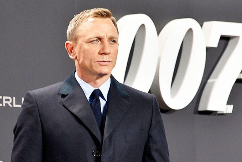 Daniel Craig, ator branco de terno escuro, em parede cinza com "007" em branco.