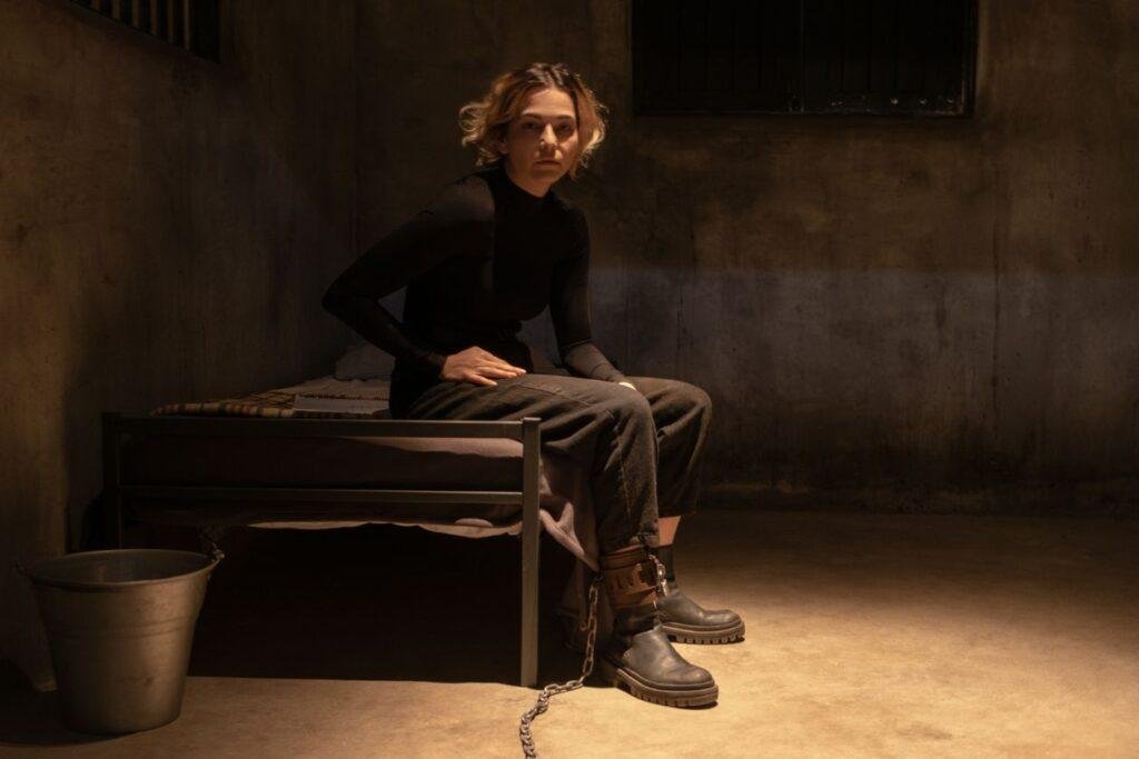 Tainá Muller como Verônica em nova temporada da série, sentada em uma cama acorrentada pelos pés.