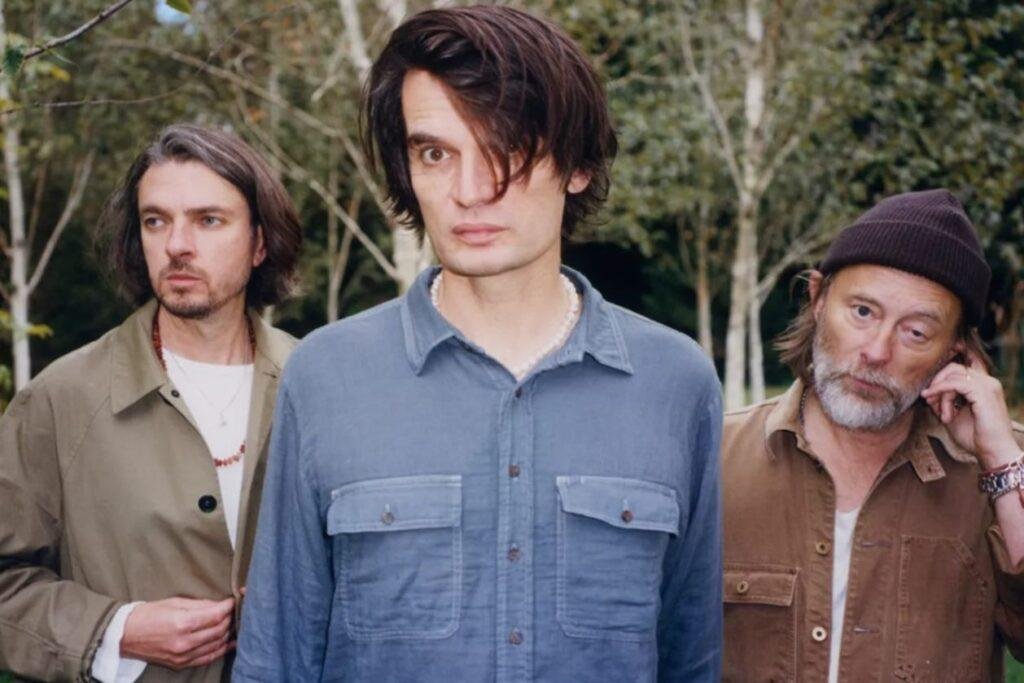 Thom Yorke, Jonny Greenwood e Tom Skinner da banda The Smile olhando para a câmera em uma floresta, com semblantes sérios/tristes