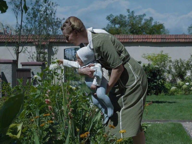 Cena de Zona de Interesse, com mulher se abaixando com criança no colo vendo plantas no jardim