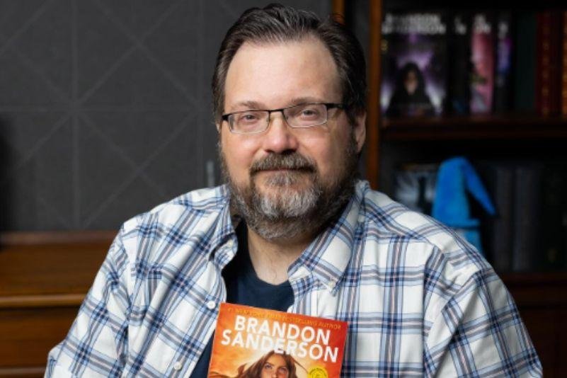 Brian Sanderson, de bluza xadrez, com o livro Defiant, de capa laranja. | Reprodução: Instagram
