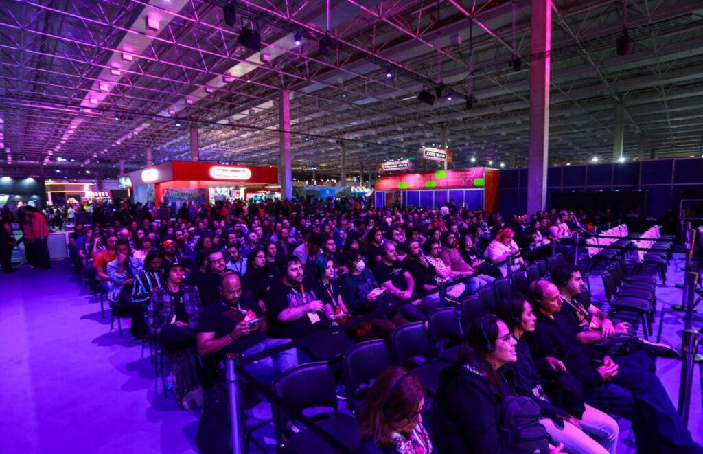 Multidão sentada em cadeiras voltadas para um palco em um evento de games, com iluminação violeta e estandes coloridos ao fundo.