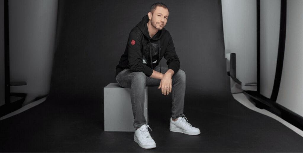 Tiago Leifert posando casualmente sentado em um bloco cinza em um cenário minimalista preto.