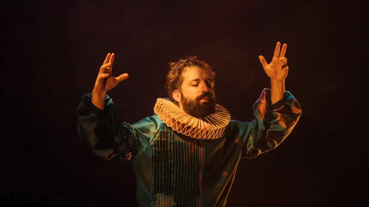 Gregório Duvivier está de olhos fechados e braços abertos durante a encenação de sua peça "O Céu da Língua". Ele veste trajes que remetem à nobreza da Idade Média. 
