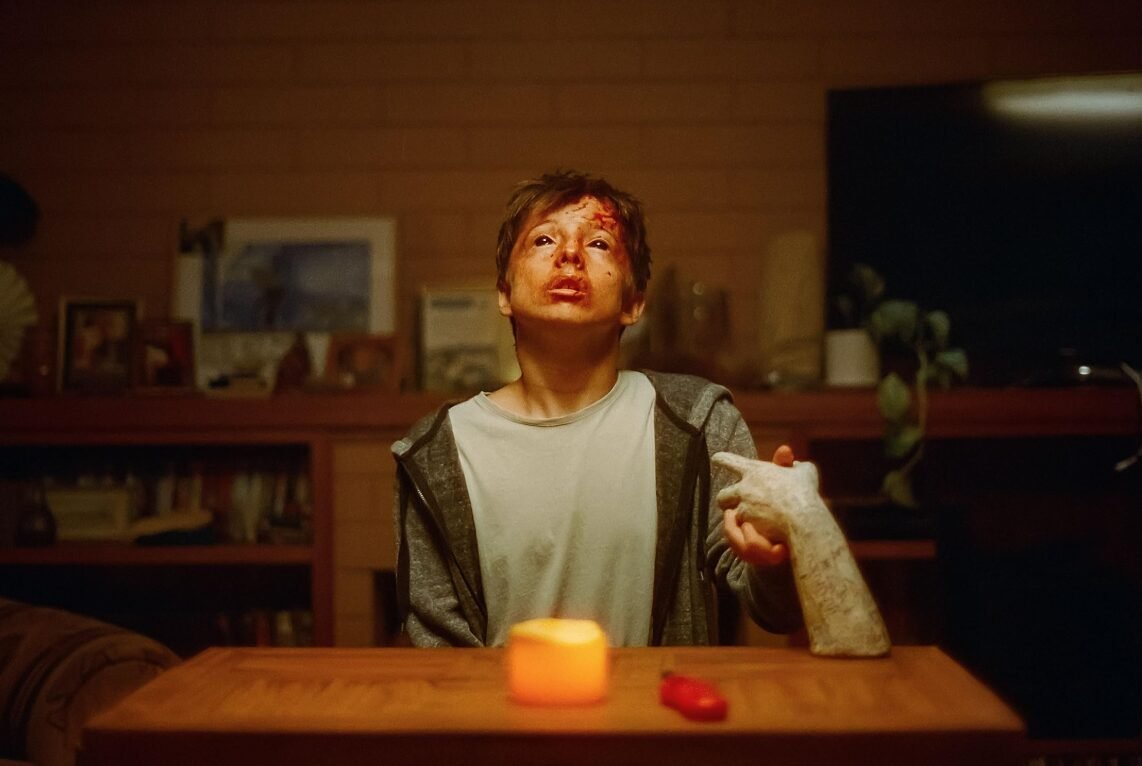 Cena do filme de terror da A24, com personagem ensanguentado segurando mão de escultura durante ritual sombrio em October A24