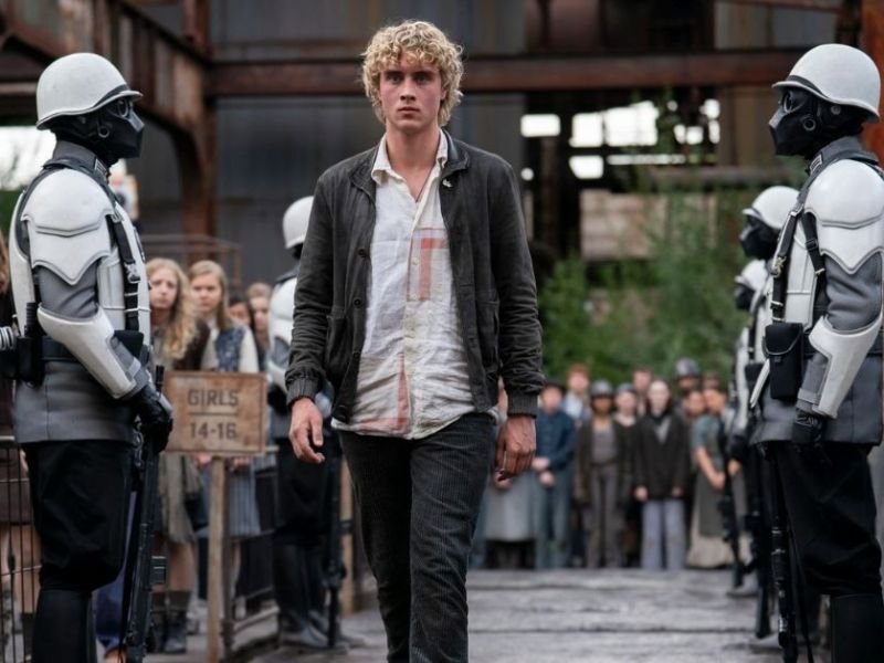 Haymitch jovem caminha entre soldados do Capitólio em cena de "Jogos Vorazes: Amanhecer na Colheita" durante a seleção dos tributos.
