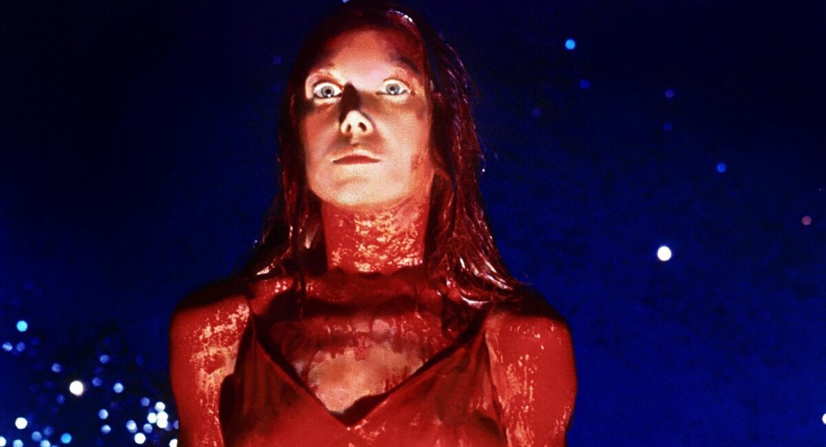 Carrie White coberta de sangue de porco, olhando para a frente com olhos arregalados e assustados.