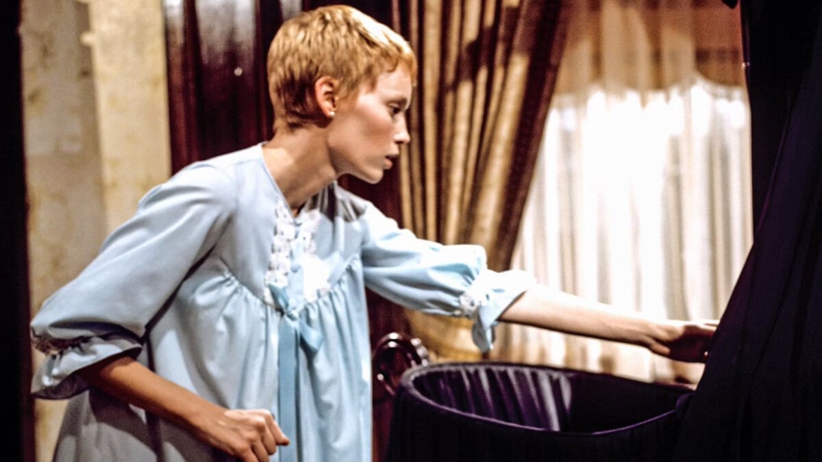 Mia Farrow, com um corte de cabelo curto, olhando para dentro de um berço escuro.
