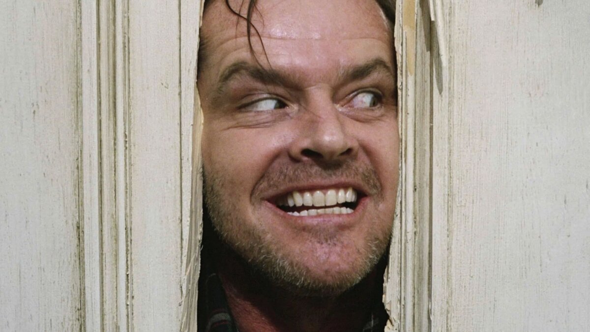Jack Nicholson com uma expressão maníaca e perturbada através de uma fresta em uma porta de madeira quebrada.