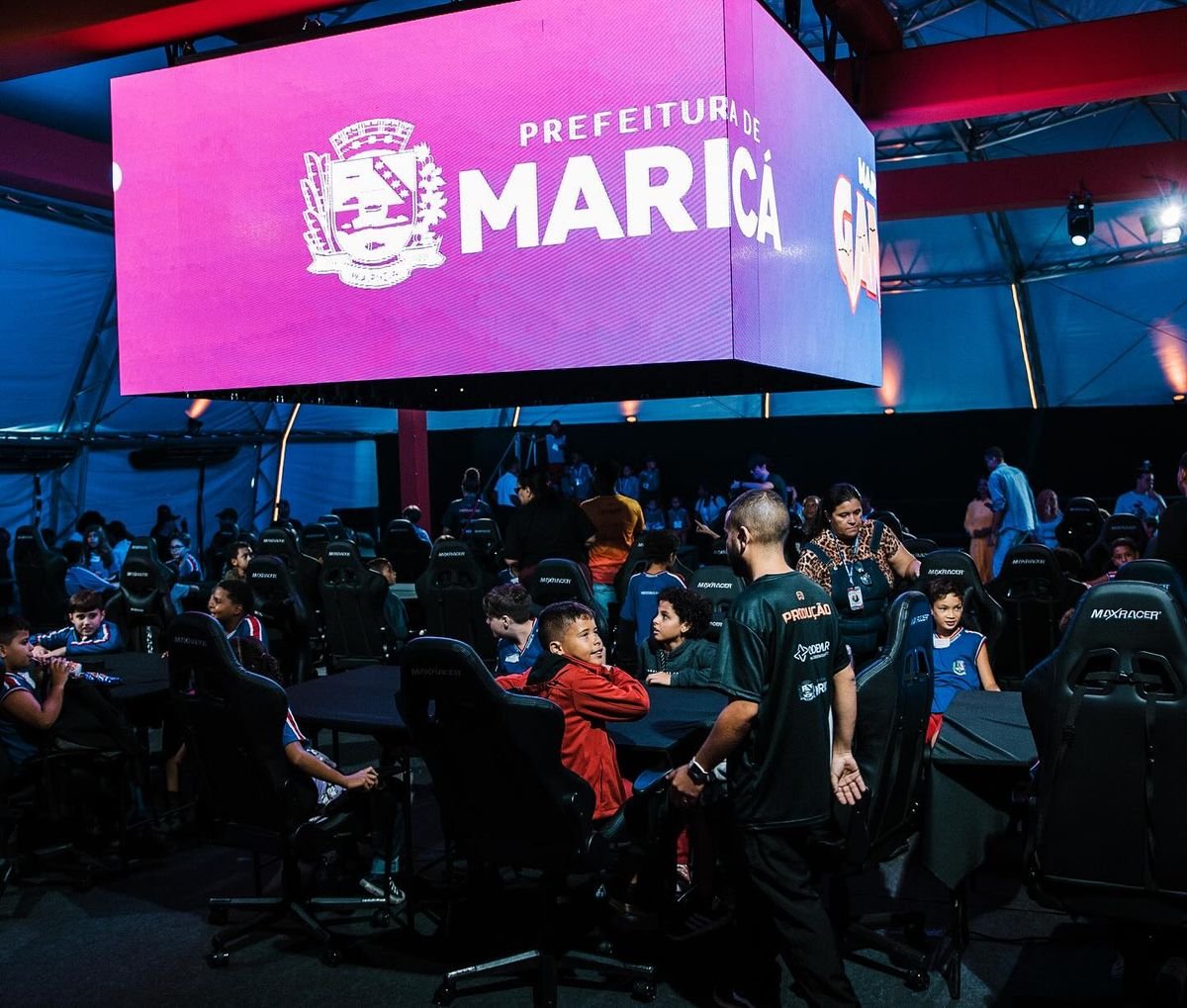 Crianças sentadas em cadeiras gamer ao redor de mesas, sob um telão suspenso com a marca da Prefeitura de Maricá.
