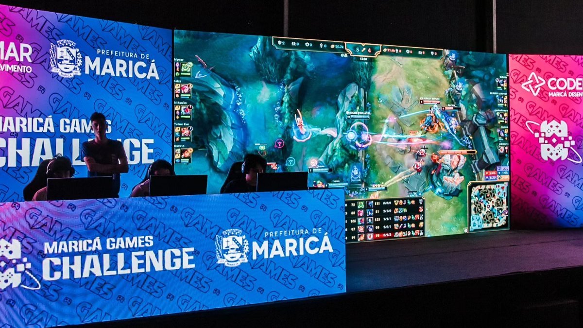 Palco com telão exibindo uma partida de League of Legends; à frente, jogadores em computadores atrás de um painel com a marca Maricá Games Challenge.
